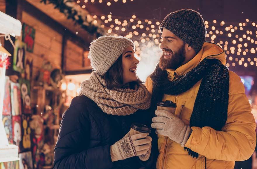 A couple wrapped up in coats and hats gaze lovingly at each other as they hold hot drinks at a Christmas market 