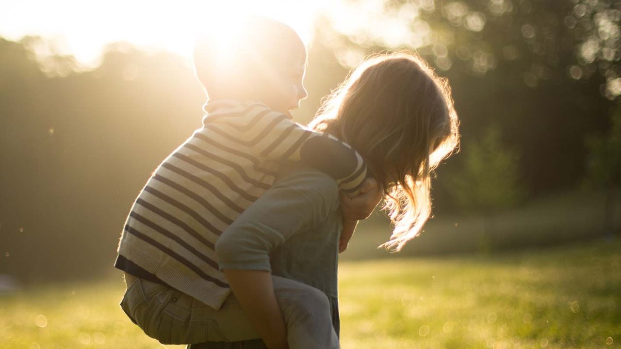 Toddler giving another child a piggyback in sunny green open space surrounded by trees