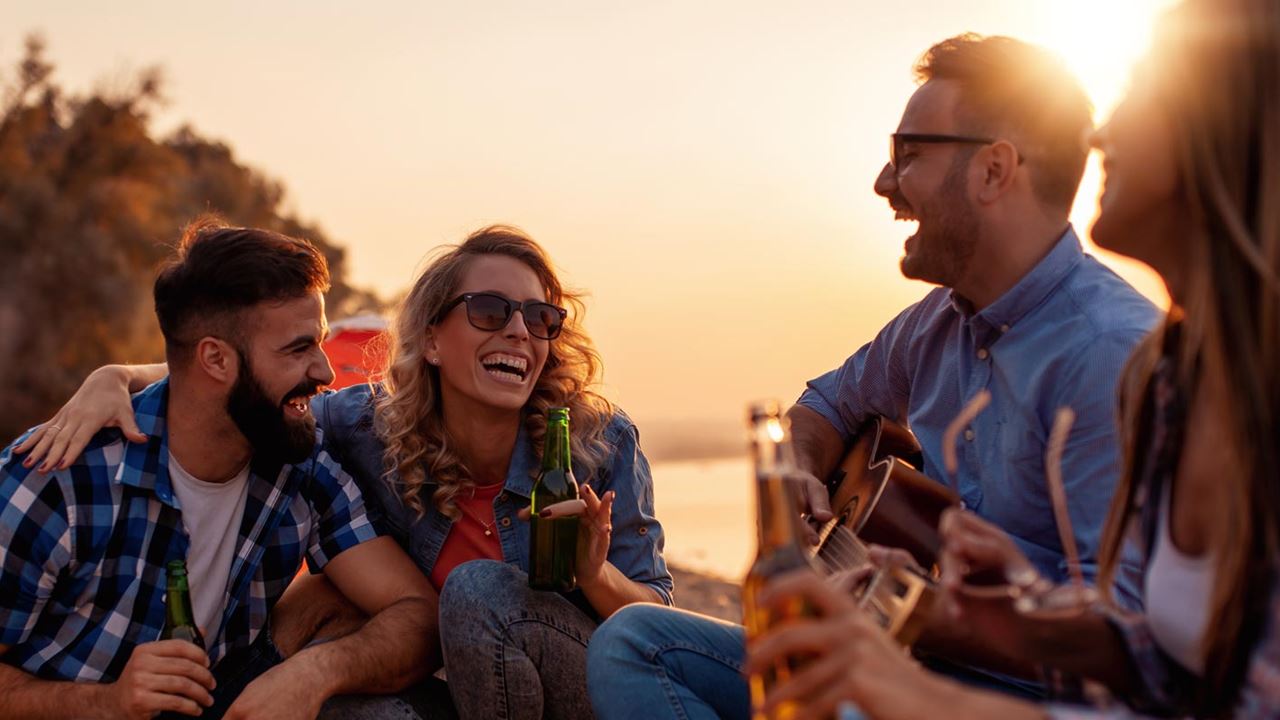 Four friends laughing with beers and guitar playing at campsite