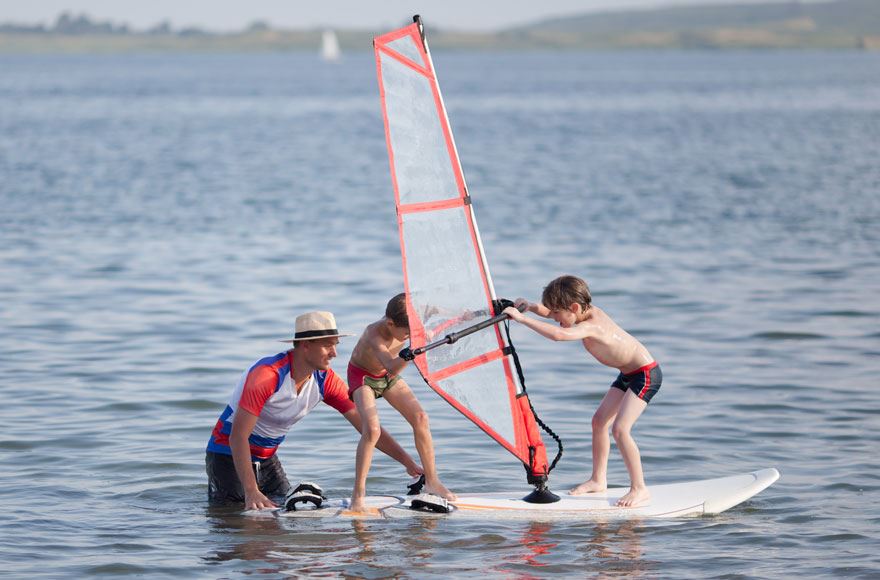 A man helping two children balance on a winder surfing board in a calm lake 