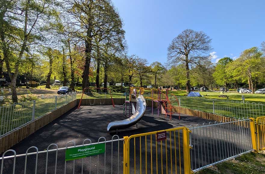 A small enclosed playground with a slide and swings with a field to the side, at Abbey Wood campsite, London