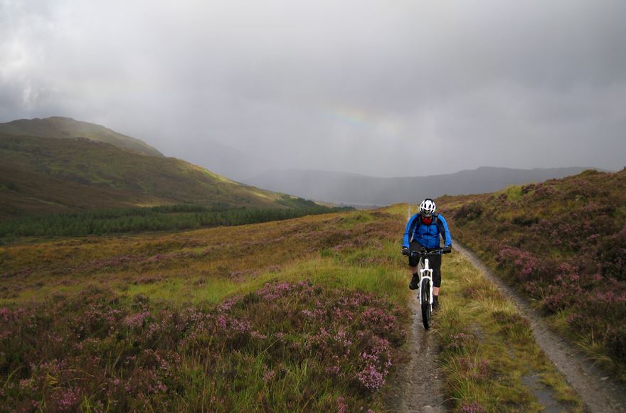 Cyclist on a white mountain  bike  wearing a blue jacket in contrast to the pale pinks, greens and browns of the moss and heathers, grey rain clouds with very faint sunbeams over the background