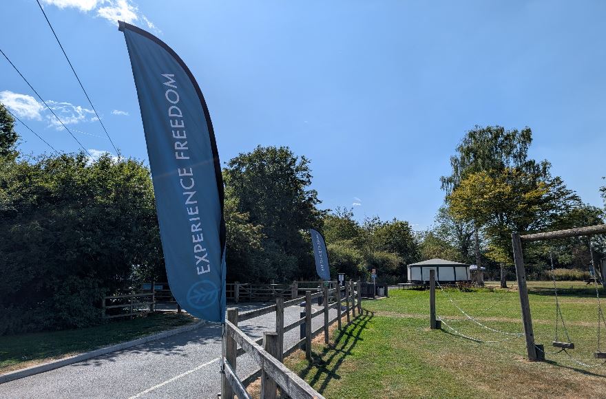 Blue Experience Freedom feather flag standing beside a gravel access path leading into the glamping field under a clear blue sky.