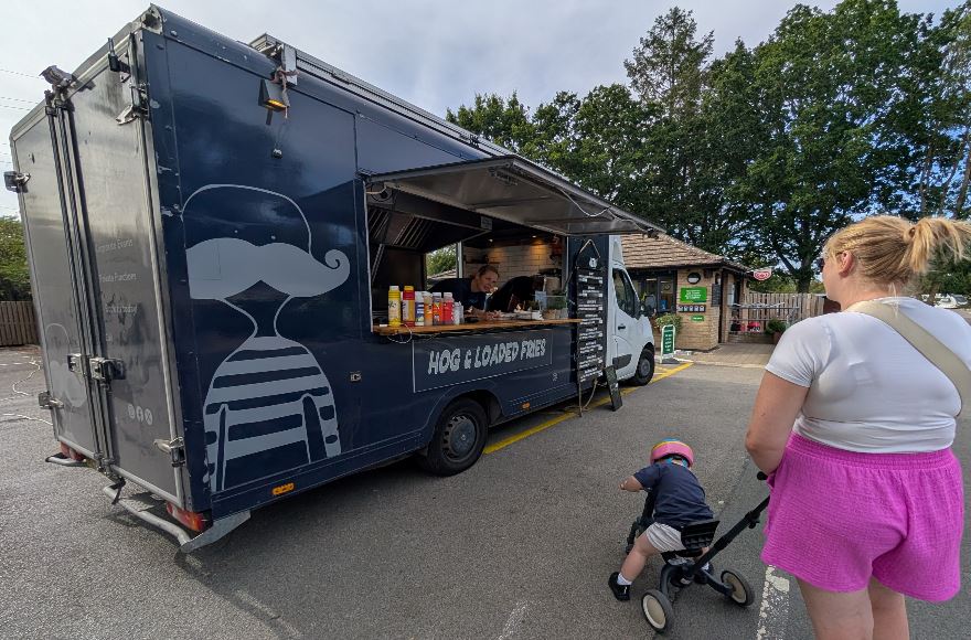 Dark grey food catering van serving hog roast and loaded fries outside the campsite reception area, with a toddler on a tricycle in the foreground.