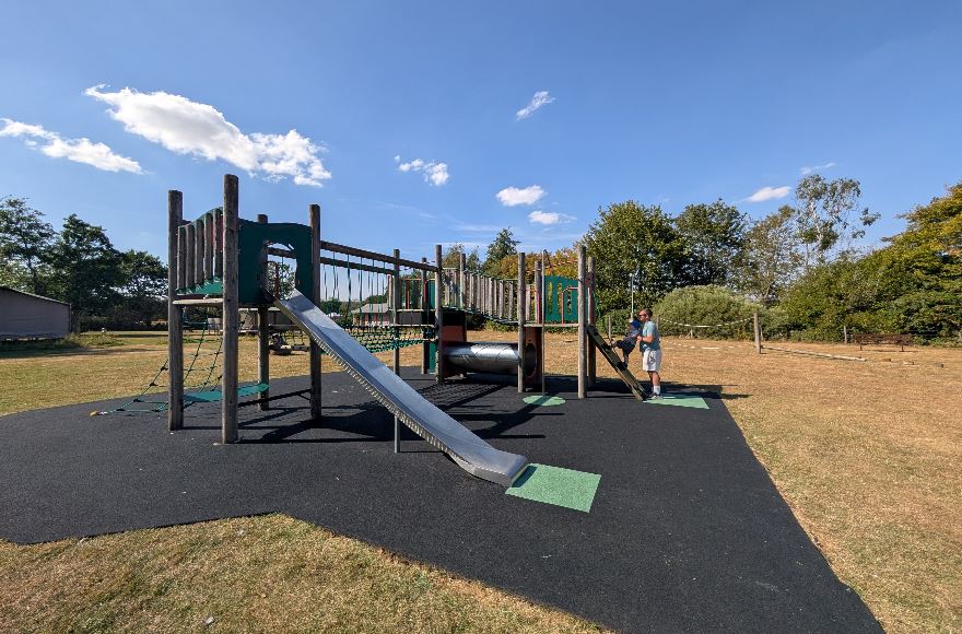 Large timber children's play structure with slides, climbing nets, and tunnels set on black safety surfacing on a sunny day.