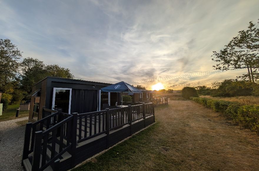 Exterior view of modern glamping pods with decked verandas facing a sunlit sky as the sun dips below the trees.