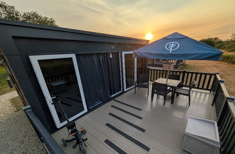 Elevated view of a glamping pod deck featuring outdoor dining table, chairs, parasol, and a toddler tricycle at sunset.