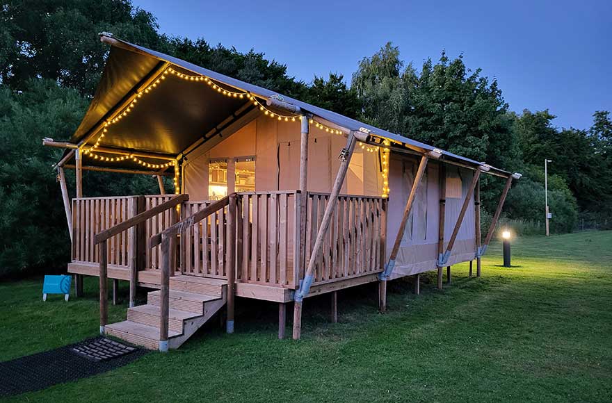 A safari tent decorated with fairy lights with a wooden deck and stairs, surrounded by nature at dusk