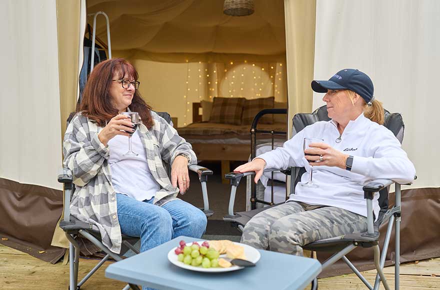 Two women relaxing in chairs and drinking wine  outside a canvas yurt