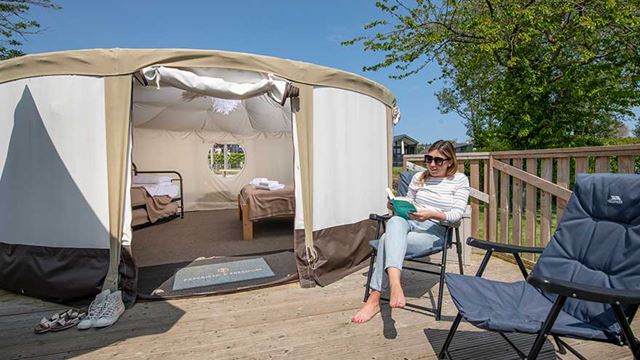 A lady relaxing in a chair outside a canvas yurt, surrounded by trees.