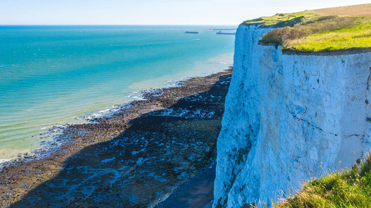 a large white chalk cliff looming over a shingle beach surrounded over a calm blue sea