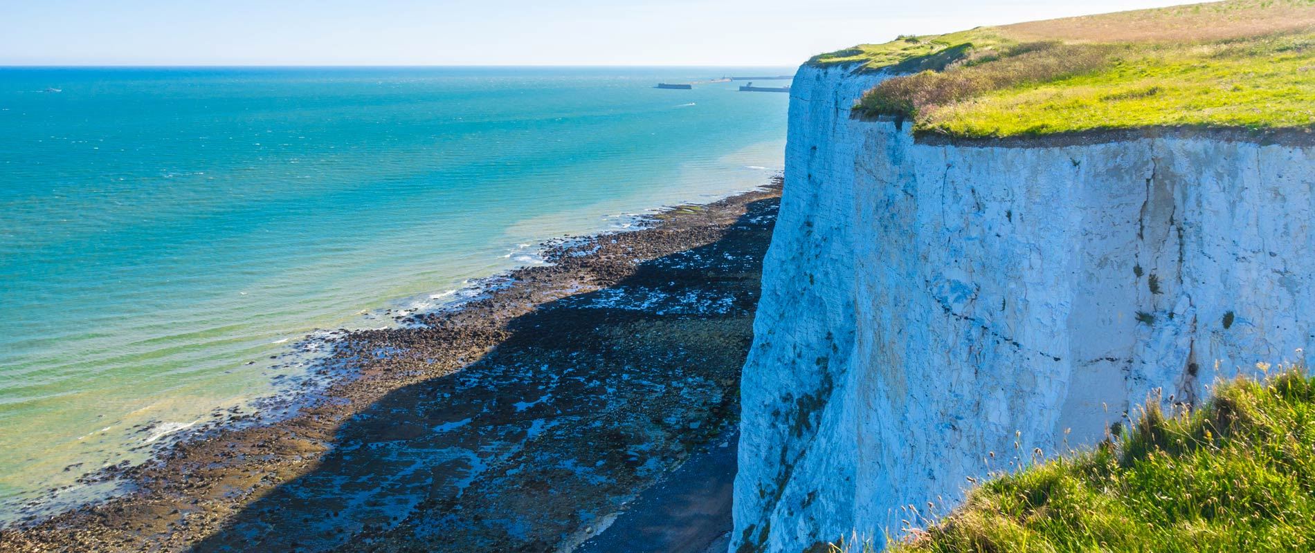 a large white chalk cliff looming over a shingle beach surrounded over a calm blue sea