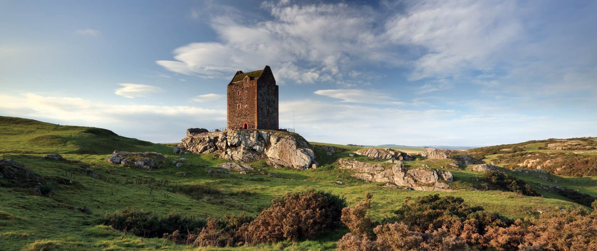 smailholm tower standing tall in a rocky and grassy field against a cloudy blue sky